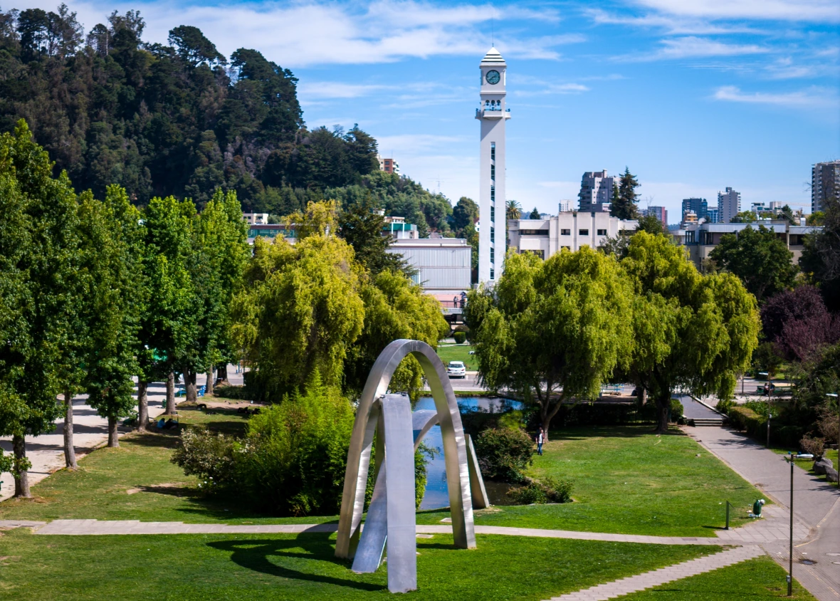 Vista panorámica del campus UdeC con escultura metálica arqueada, laguna, torre del reloj y áreas verdes.
