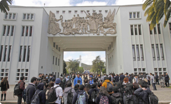 Arco universitario blanco con relieve escultórico; gran grupo de estudiantes con mochilas se congrega debajo.