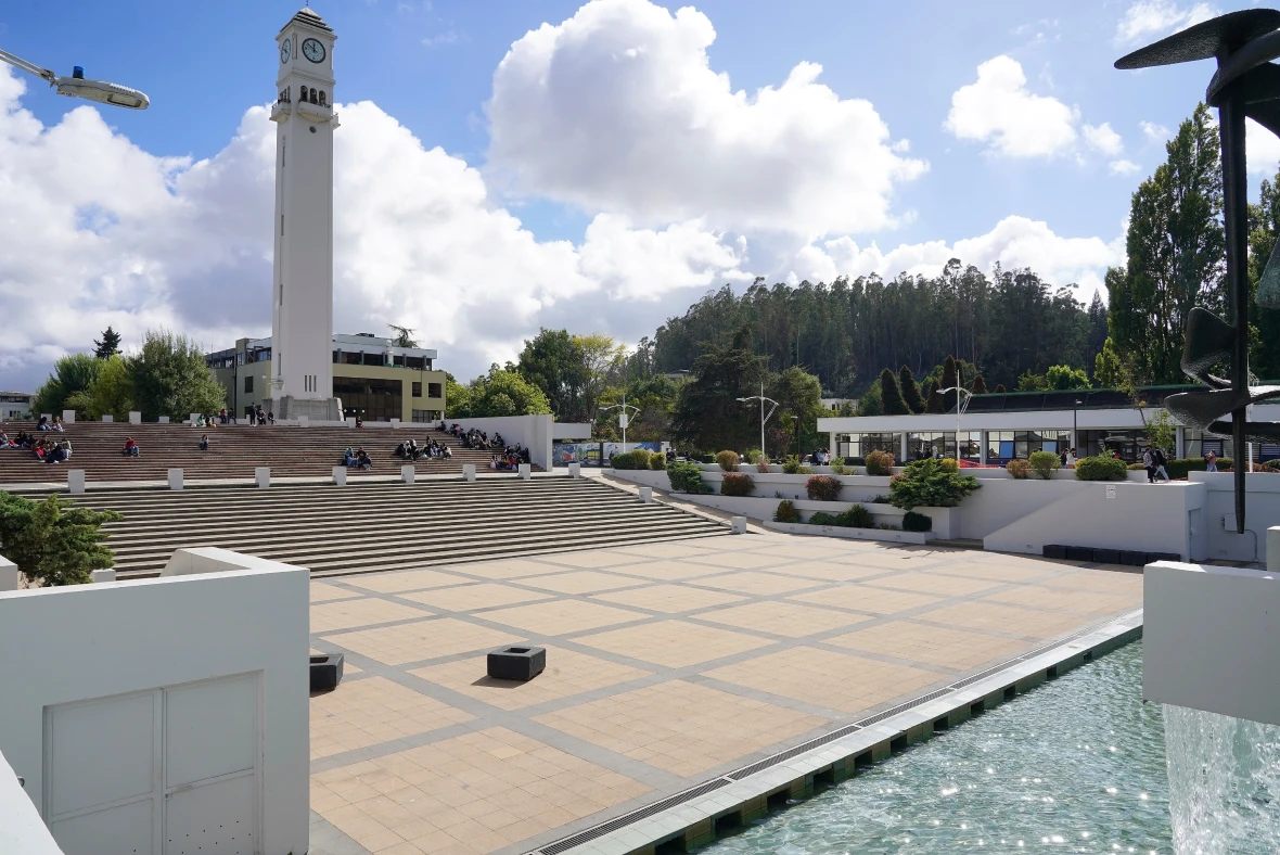 Campanil UdeC frente a amplia plaza con escaleras, estudiantes sentados y fuente lateral, bajo cielo nublado parcial.