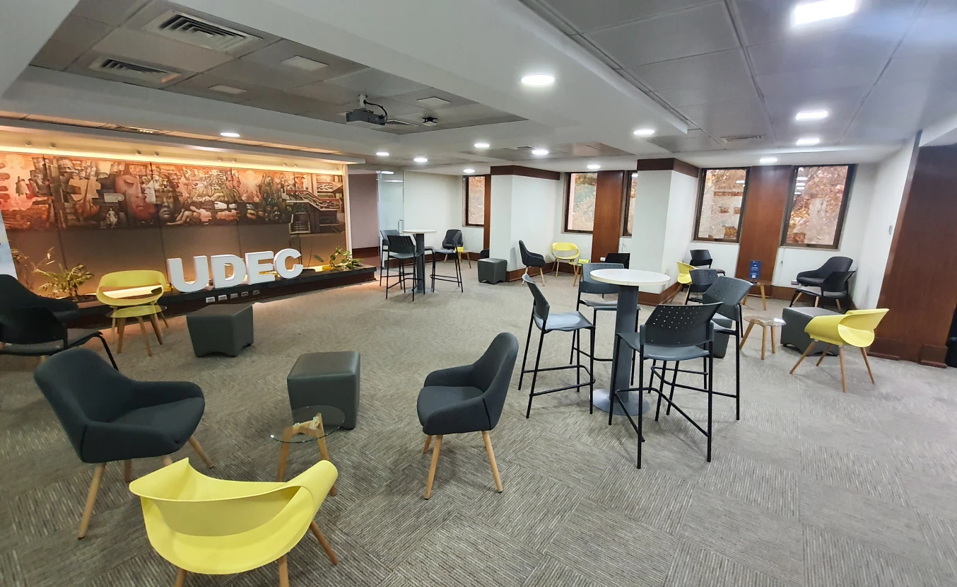 Espacio interior del campus UdeC Santiago, sala lounge con sillones amarillos y grises, mesas altas y mural iluminado.
