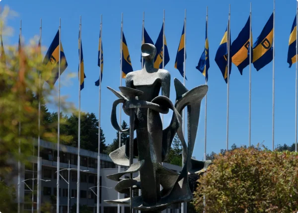 Escultura abstracta ante una fila de banderas azul - amarillo de la UdeC, con un edificio y árboles al fondo.