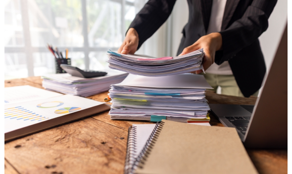 Persona con blazer organiza pilas de documentos con separadores sobre escritorio de madera, junto a laptop y gráficos.