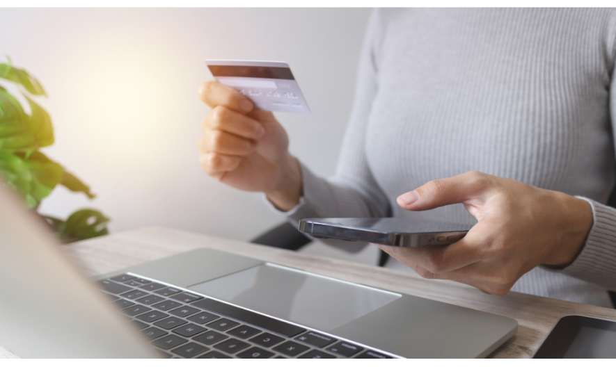 Persona con suéter gris realiza pago online sosteniendo tarjeta y smartphone frente a laptop sobre escritorio.