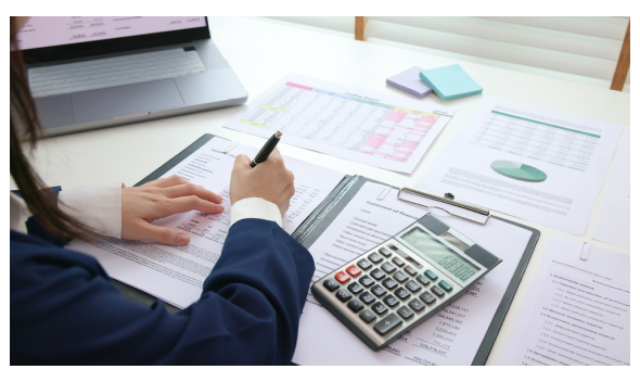 Persona analiza documentos financieros en escritorio con calculadora, laptop y hojas con tablas y gráfico circular.