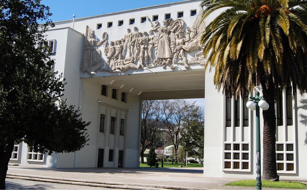 Pórtico universitario blanco con gran relieve en la parte superior, al costado palmera en día soleado.