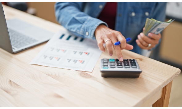 Persona con chaqueta mezclilla teclea en calculadora mientras sostiene billetes; laptop y hoja con gráficos sobre mesa. Representa la Unidad de Remuneraciones.