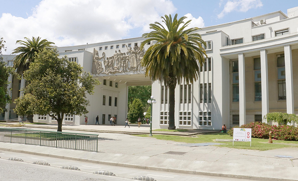 Fachada blanca con arco escultórico de la UdeC, arboles y columnas laterales.